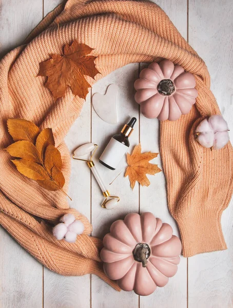 Autumn Skin Care products - mockup bottle of cosmetics, jade massager, guasha, autumn leaves, pumpkins, knitted sweater. Seasonal beauty routine and organic skin care concept.