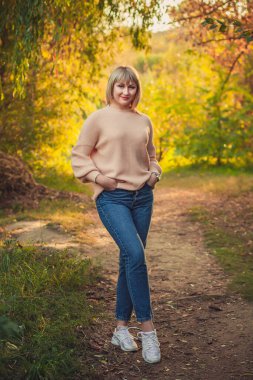 Kısa saçlı sarışın bir kadın örülü bir kazakla ormanda yürüyor. Sonbahar ormanlarında doğa yürüyüşü..