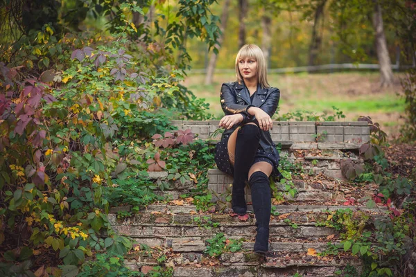 Young woman with bob haircut and fringe in a black leather jacket and high black jackboots is sitting on an abandoned staircase in autumn. Staged photo shoot