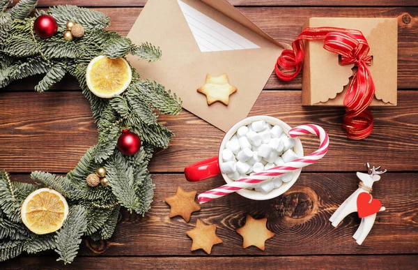 Vánoční věnec, dárek, kávový hrnek s marshmallows, obálka a domácí sušenky a různé vánoční ozdoby na dřevěném stole. Rustikální vánoční věnec, plochý ležel. — Stock fotografie