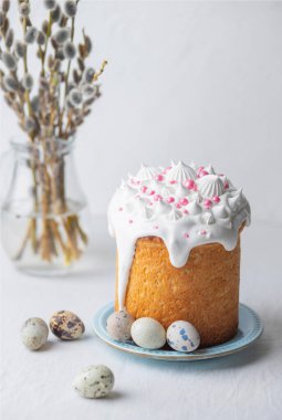 Traditional homemade Easter cake, decorated with glaze and pastry sprinkles on a blue plate on a white table with quail eggs and willow branches in the background. Happy Easter. Vertical photo. Front