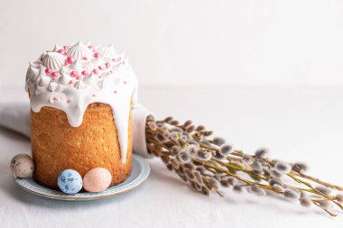  Traditional homemade Easter cake, decorated with glaze and pastry sprinkles on a blue plate on a white table with colored quail eggs and willow branches in the background. Happy Easter. 