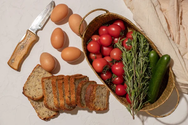 Färska tomater, gurkor, några kvistar rosmarin i en korg, hönsägg och skivat bröd på en vit yta. Skördar. Jordbruksprodukt. Ovanifrån Hård skugga. — Stockfoto