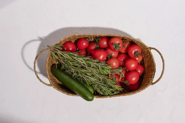 Pomodori freschi, cetrioli e qualche rametto di rosmarino in un cestino di vimini su una superficie bianca. Vendemmia. Prodotto agricolo. Ombre dure. — Foto Stock