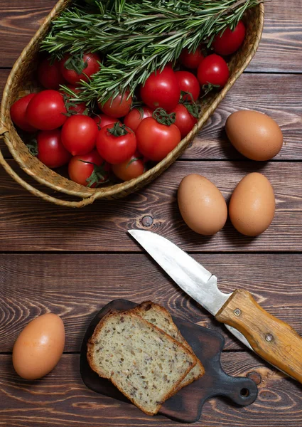 Färska tomater, gurkor, några kvistar rosmarin i en korg, hönsägg och skivat bröd på ett träbord. Skördar. Jordbruksprodukt. — Stockfoto