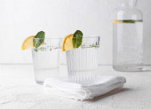 Dvě sklenice čerstvé vody, zdobené citronovými klíny a mátovými listy na bílém stole. Selektivní soustředění. Pohled zepředu