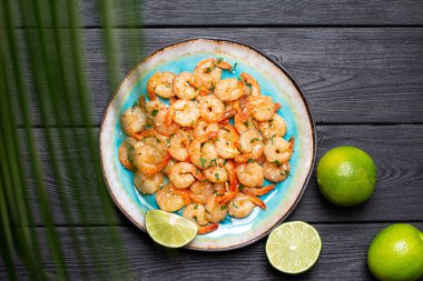 Mavi porselen bir tabakta sarımsaklı karidesler ve dilimlenmiş limonlar siyah ahşap bir masada. Üst görünüm
