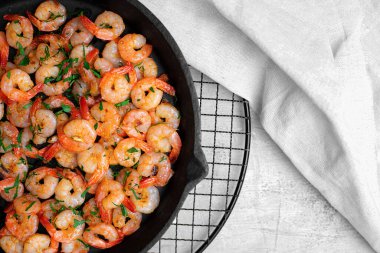 Beyaz keten peçeteyle beyaz bir masada, dökme demir tavada sarımsaklı karidesler. Üst görünüm