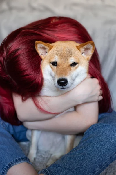 Μια γυναίκα με κόκκινα μαλλιά αγκαλιάζει σφιχτά ένα χαριτωμένο κόκκινο σκυλί Shiba Inu, κάθεται στην αγκαλιά της στο σπίτι. Κοντινό πλάνο. Εμπιστοσύνη, ηρεμία, φροντίδα, φιλία, η έννοια της αγάπης. Χαρούμενες στιγμές της ζωής. — Φωτογραφία Αρχείου