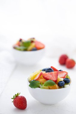 Taze çilekli taze meyve salatası, kivi, kayısı ve böğürtlen, beyaz bir masada taze nane yapraklarıyla süslenmiş.