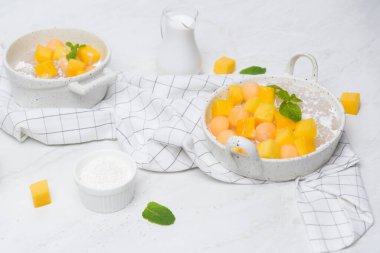 Tayland tatlıları: Hindistan cevizi sütüyle ev yapımı sago ve beyaz bir kasede kareler ve kavun topu şeklinde kesilmiş mango ve bir masa örtüsü ekoseli mermer bir masaya yerleştirilmiş..