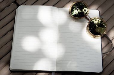 Boş satır defteri el yazısı. Bulanık güneş gözlükleriyle dışarıdaki bambu ahşap bir masa üzerinde. Gölge ve güneş ışığıyla süslenmiş.