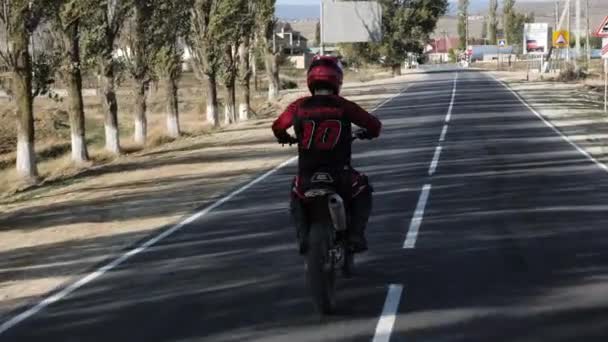 Motociclista en velocidades de casco a lo largo de camino de asfalto al pueblo — Vídeo de stock