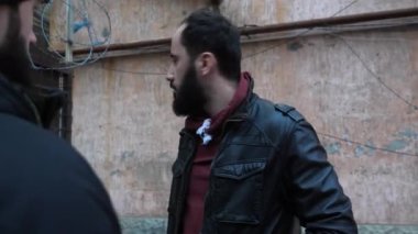 Ceketli sakallı adam etrafına bakar ve keşle konuşur.