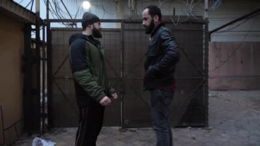 Siyah şapkalı sakallı adam deri ceketli adamla konuşuyor.