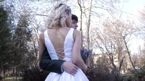 Groom hugs pretty bride in stylish wedding dress in park — Stock Video