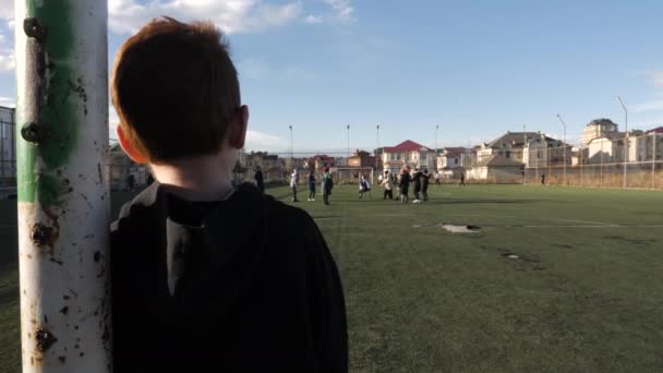 Liten pojke tittar på barn som spelar fotboll på gröna fältet Stockfilm