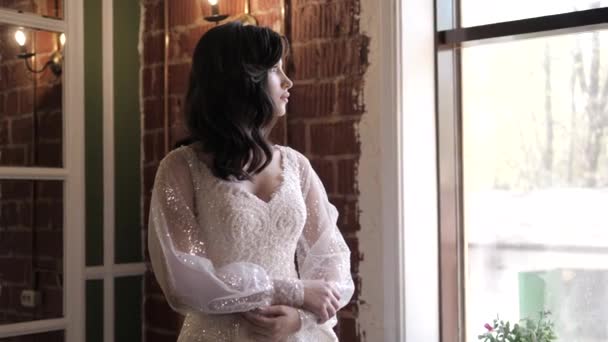 Brunette dame en robe de mariée glamour regarde par la fenêtre — Video