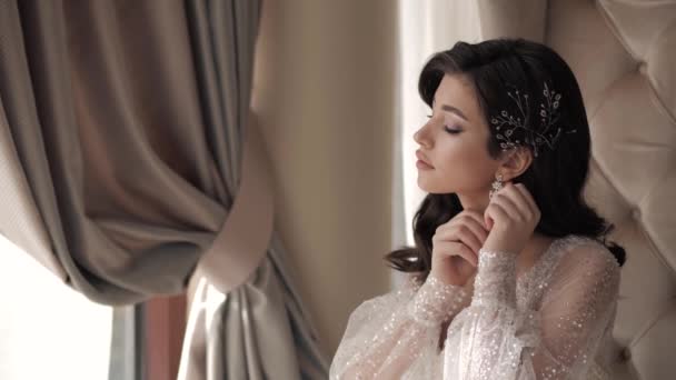 Drömmande brud i glänsande klänning justerar örhänge nära fönstret Stockvideo