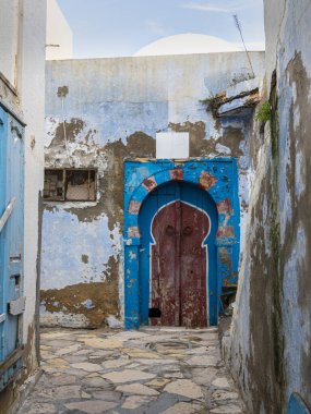 Hammamet Medine 'nin antik ve dar sokaklarında derin derin bir yolculuk. Yumuşak, bulutlu bir kış gökyüzünün altında, ikonik beyaz yıkanmış duvarlar ve canlı mavi kapılar, otantik, q 'nun mükemmel bir şekilde sergilendiği, dingin ve sessiz bir renk paleti oluşturur.