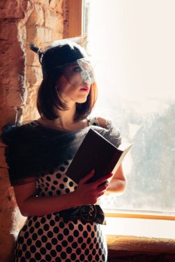 Geçen yirmili yılların tarzında giyinmiş siyah şapkalı, duvağı açık omuzları açık, elinde kitapla pencerenin kenarında duran bir kız..