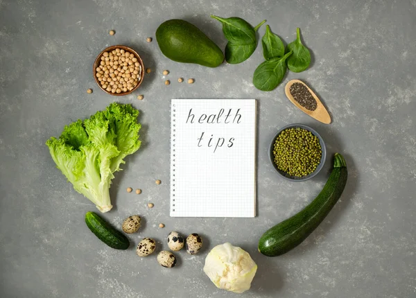 Diet plan with greens and fresh vegetables in a paper notebook on a gray table surface.