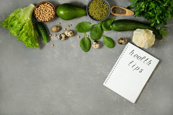 Diet plan with greens and fresh vegetables in a paper notebook on a gray table surface.