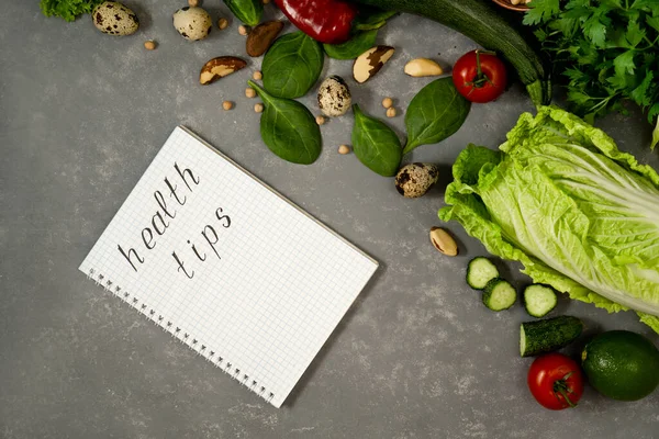 Diet plan with greens and fresh vegetables in a paper notebook on a gray table surface.