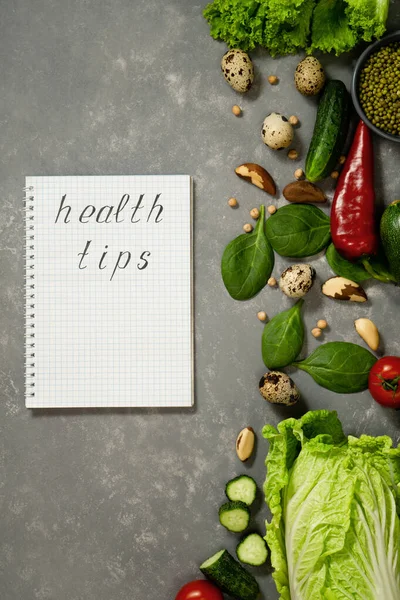 Diet plan with greens and fresh vegetables in a paper notebook on a gray table surface.