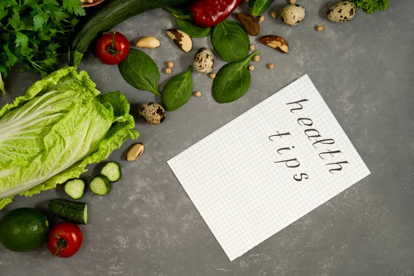 Diet plan with greens and fresh vegetables in a paper notebook on a gray table surface. Words Health tips are written on a notebook