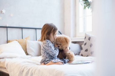 Oda yeni yıl için dekore edilmiş, bir kız bir ayıyla yatakta oturuyor, pelüş bir oyuncak okulda oynuyor, kitap okuyor, sayfaları karıştırıyor, bir anaokulu öğrencisi.
