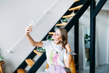 Avrupa görünümlü bir kız telefonla, sosyal ağlarla, uzaklıkla, uzaktan çalışma ve iletişimle, evin ve ofisin içinde kendi fotoğraflarını çekiyor.