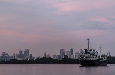 Bangkok, Tayland - 04 Ocak 2021: Akşam Chao Phraya Nehri 'nin ortasına park etmiş bir kargo gemisi ve arkasında da yeşil ormanlar ve gökdelenler var. Seçici odak.