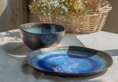 Beyaz desenli masa örtüsünün üzerinde dokuma sepetinde seramik bulaşık takımı ve bir buket çiçek. Seramik tablo takımı, Seçici odak.