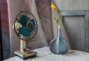 Bangkok, Tayland - Şubat 06.2021: Mavi ahşap vazodaki sarı çiçek ve eski beton duvarlı beyaz desenli masa örtüsü üzerindeki vintage fan. Ev dekorasyonu, Seçici odak.
