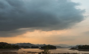 Gün doğumunda veya günbatımında arkaplanda dağlar olan göl kıyısı manzarası. Tam olarak odaklanamıyorum..
