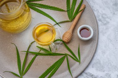 Rahatlatıcı ve keyifli bir fincan kenevir bitkisel çay ve yeşil marihuana yapraklı kök mermer masa üzerinde bal ile uyku ve endişe için tıbbi amaçlar. Bitki çayı ve sağlıklı içecek konsepti. Boşluğu kopyala, Seçici odak.