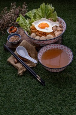 Domuz etli erişte, kıymalı domuz eti, sahanda yumurta, sebze, kase içinde çorba ve kaşık ve kereste arkaplanlı çubuk. Tayland eriştesi çorbası.