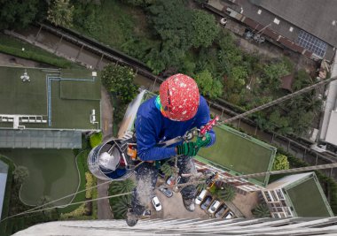 Bangkok, Tayland - 3 Kasım 2025 - güvenlik kaskı ve emniyet kemeri takan bir işçi, yüksek bir binanın dış tarafındaki iplerle asılmış. İşçi binanın ön cephesindeki eski boyaları kazıyacak..