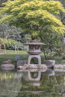 Londra, İngiltere - 27 Nisan 2025 - Hollanda parkındaki Kyoto bahçesinin arka planında yemyeşil bitkiler bulunan zen gölü suyu üzerindeki Japon taş lambasının çarpıcı görüntüsü. Londra 'da sakin Japon peyzajında dekoratif taş fener, yazı için alan.