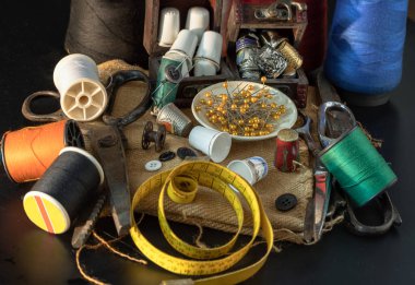 Çeşitli renklerde iplik makaraları, küçük ahşap hazine sandığında yüksükler, çelik makas, yeşil minyatür makaralara sıkışmış iğneler, iplik ve mezura şeritleri, koyu arkaplandaki küçük yuvarlak tepside bulunan iğneler. Dikiş ögeleri seti ya da Dikiş