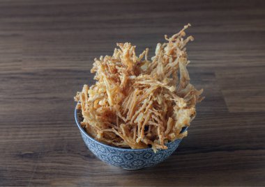 Kızarmış enoki, ayrıca bol yağda kızartılmış iğne mantarı olarak da bilinir, baharatlı un ile kaplanır ve altın-kahverengi, kıtır kıtır bir dokuda kızartılır; ahşap arka planda mavi ve beyaz desenli bir kasede servis edilir ve popüler bir lezzet türüdür.