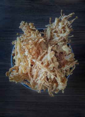 Kızarmış enoki, ayrıca bol yağda kızartılmış iğne mantarı olarak da bilinir, baharatlı un ile kaplanır ve altın-kahverengi, kıtır kıtır bir dokuda kızartılır; ahşap arka planda mavi ve beyaz desenli bir kasede servis edilir ve popüler bir lezzet türüdür.