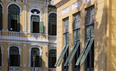 Parşömenin Neoklasik derinliğini vurgulayarak ılık, açık renkli cephede keskin gölgeler oynar. Mimari dokunun ortasında koyu yeşil panjurlu açık bir pencere güneşe karşı cesur ve zarif bir odak noktası olarak duruyor..