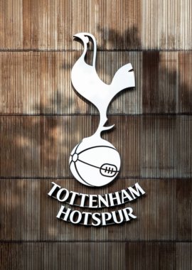 Benekli güneş ışığı, Tottenham Hotspur simgesi boyunca yumuşak gölgeler bırakır ve bu gölge dikey ahşap kalasların dokulu arka planına monte edilir. Amblemler pürüzsüz, kıvrımlı siluet ve sivri gagalı horoz duruşu gururla bağcıklı futbol topunun üstünde durur.