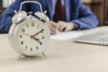 Ofis masasında klasik çalar saat. Bulanık arka planda, patron belgeleri imzalar. Ofis çalışanı. Zaman kavramı