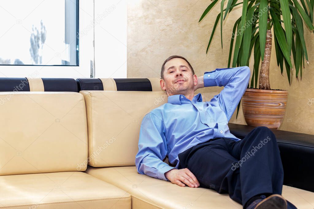 The concept of the need for rest during work. A tired manager in a blue shirt is lying on the couch in his modern office