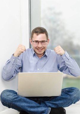 İşadamı, mavi gömlekli bir ofis çalışanı, pencere eşiğinde dizüstü bilgisayarla oturuyor. Duygusal olarak internetten iletişim kuruyor. Çevrimiçi iletişim. Mutlu duygular. Dikey resim