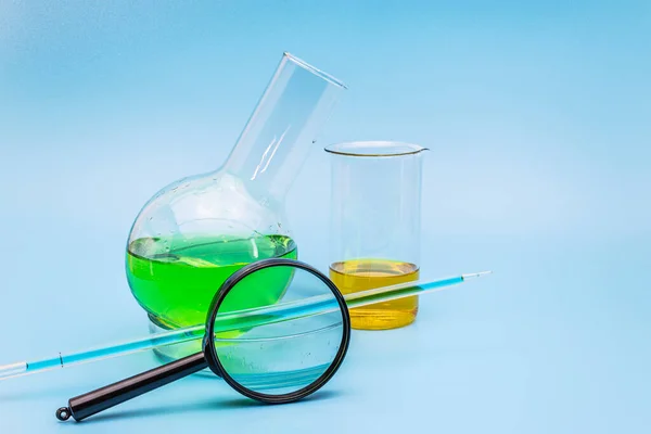 ガラス実験室のフラスコとビーカーは 優しい青色の医学的背景に拡大ガラスと緑と黄色の溶液をカップに入れます 医学と化学の概念は分析する コピースペース — ストック写真