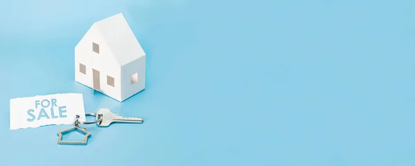 The concept of mortgage, sale and rental of housing and real estate. Buying a home. A mock-up of a paper house with keys on a blue background. Biroka - for sale. Banner format. Copy space